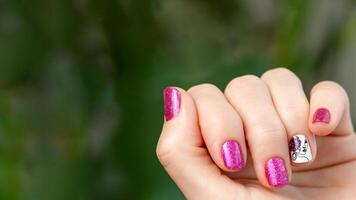 Female hand with pink manicure. Woman fingernails with pink holographic nail polish lacquer on green lush background. Aesthetic visual of red violet manicure with lady silhouette stamping. Copy space photo