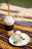 Homemade fresh strawberry cheese cake with lemon piece in white dish and Ice latte shake coffee with whipped cream in plastic glass on Thai native pattern tablecloth in natural sunlight in cafe photo