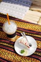 Fresh strawberry cheese cake with lemon piece in white dish and Ice latte shake coffee with whipped cream in plastic glass on Thai native pattern tablecloth in natural sunlight in cafe photo