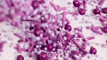 Vibrant purple and pink candy sprinkles cascade onto a surface, creating a textured, colorful pattern in macro view video