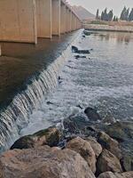 a wide waterfall flows beneath a bridge in the center of a wadi. photo