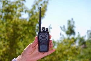 Handheld walkie talkie being held against a sunny outdoor background with greenery photo