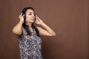 Indian female individual looking sideways, touching her headphones and listening intently to a podcast. Focused woman using wireless headset, standing isolated over brown background in studio. photo