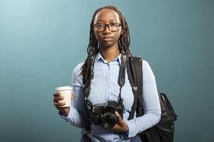 Young woman poses in studio, dressed casually with dslr camera hanging on her neck and coffee in hand. African american female model shows a focused yet professional photographer look. photo