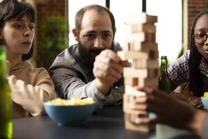 Bearded caucasian man with focused expression, carefully removing block from wooden tower during indoor game session. Male individual seated with female friends playing tower stacking game. photo