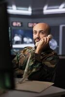 Military officer looking at surveillance footage, analyzing data to allocate resources to units based on mission needs, making telephone call. Agent examining data on screen and talking on phone photo