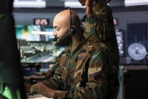 Army dispatcher looking at surveillance transmission on monitors, relaying data to troops using headphones microphone. Military operative transmitting reconnaissance info photo
