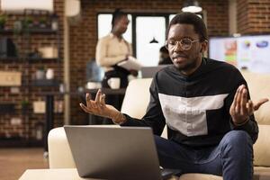 Focused businesswoman with earbuds, seated on cozy sofa, participating in virtual meeting in brick wall office. African american male manager using laptop for videoconference with remote startup team. photo