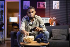 Online gamer enjoying shooter gaming competition with popcorn at home, participating in multiplayer games tournament with friends during game night. Young adult having fun with championship. photo
