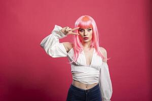 Trendy young woman with pink hair shows a peace sign and puckers lips playfully for the camera. Stylish female model poses confidently against pink background, expressing carefree vibes and charm. photo
