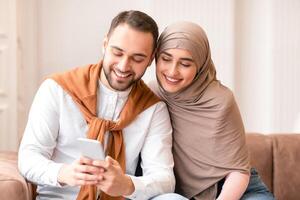 Front View Of Muslim Couple Using Cellphone Sitting On Sofa Together At Home. Middle Eastern Spouses Browsing Internet And Using Social Media On Smartphone Indoors. New Mobile Application Concept photo