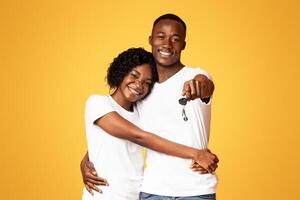 Happy young black couple with new car keys hugging and smiling over orange studio background, copy space. Cheerful african american man and woman embracing and showing key from auto photo