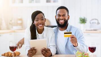 Portraif Of Happy Black Couple With Digital Tablet And Credit Card In Kitchen, Young African Spouses Making Food Or Groceries Delivery Order, Sitting At Table With Red Wine Glasses, Smiling At Camera photo