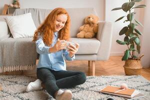 Mobile Gaming Concept. Excited Red-Haired Teenage Girl Using Smartphone, Playing Online, Sitting On The Floor Carpet And Leaning On The Sofa At Home In Living Room Or Bedroom. Copy Space photo