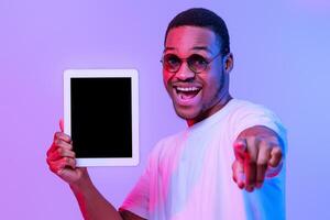 App For You. Joyful African American Guy Holding Digital Tablet With Black Screen And Pointing At Camera, Cheerful Millennial Man Standing Under Neon Light Over Purple Studio Background, Mockup Image photo