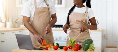 Black Millennial Couple Cooking In Kitchen Together And Using Laptop Computer, Cheerful Young Spouses Wearing Aprons, Preparing Healthy Food For Dinner At Home With Recipe From Internet, Copy Space photo