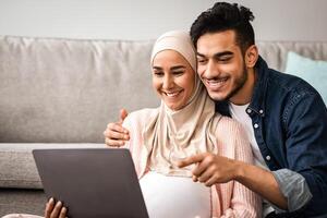 Cheerful Pregnant Muslim Woman In Hijab And Her Husband Using Laptop At Home Together, Middle Eastern Parents Shopping Online With Computer Or Watching Movie, Man Pointing At Screen, Closeup photo