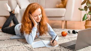 Distance Education. Smiling schoolgirl lying on the floor carpet with her laptop and writing in notebook. Girl watching tutorial, lecture or webinar, studying online at home. E-learning photo