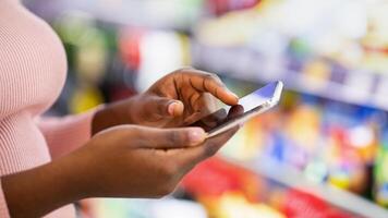 Cropped view of young black woman checking grocery list app on cellphone at supermarket, copy space. Closeup of millennial African American lady with mobile device shopping for food at store photo