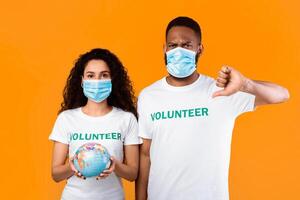 Two Mixed Volunteers Wearing Face Masks Holding Earth Globe And Gesturing Thumbs Down Standing Over Yellow Studio Background. Negative Influence Of Covid-19 Pandemic On Ecology Concept photo