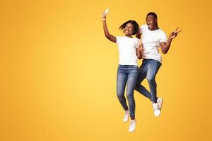 Love picture. Happy african-american couple in love making selfie on smartphone while jumping in the air, orange studio background, empty space. Showing peace gesture, full length shot photo