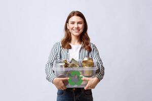 Waste Recycle. Smiling Young Woman Carrying Container Filled With Tin Cans, Millennial Eco Volunteer Lady Enjoying Garbage Recycling, Standing Over Light Gray Background In Studio, Free Space photo