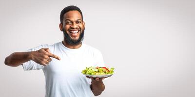 Eat Healthy. Cheerful Black Guy Pointing Finger At Plate With Vegetable Salad Dieting Standing Over White Studio Background. Male Nutrition And Weight Loss, Slimming Concept photo