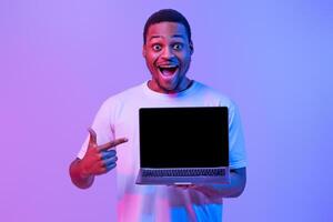 Check This. Overjoyed Black Guy Pointing At Laptop Computer With Blank Screen, Demonstrating Free Copy Place For Your Website Design, Standing In Neon Purple Lighting, Mockup Image photo