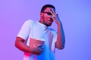 Scary Movie. Thrilled Black Guy In 3D Glasses Watching Horror Film, Millennial African American Man Holding Bucket With Popcorn And Covering Eyes With Afraid, Standing In Neon Purple Light, Free Space photo