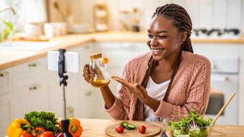 Culinary Vlog. Smiling Black Food Blogger Woman Recording Content In Kitchen, Using Smartphone On Tripod For Filming Healthy Salad Recipe, Demonstrating Jug With Olive Oil At Camera, Free Space photo