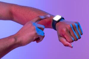 Unrecognizable african american male using smartwatch with blank screen, checking digital data, black guy standing in neon light over purple studio background, cropped image with copy space, mockup photo