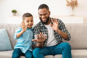 African American Dad And Son Calling Via Phone Waving Hello To Smartphone Screen Sitting On Couch At Home. Father And Kid Boy Communicating Online Indoor. Distant Communication. Selective Focus photo