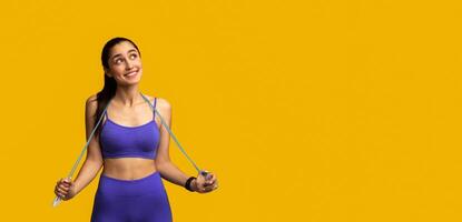 Sports Lifestyle And Weight Loss. Portrait Of Excited Young Lady Holding Jumping Rope Around Neck, Standing At Studio And Looking Away At Free Space, Isolated On Orange Background. Advertisement photo