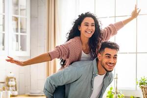 Joyful Middle Eastern Spouses Having Fun Together In Kitchen Interior, Handsome Arab Man Piggybacking His Cheerful Wife, Happy Attractive Lady Enjoying Ride, Spreading Hands Like Flying, Copy Space photo