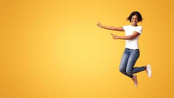 Emotional african american millennial lady smiling and pointing at advertising or announcement. Excited black woman jumping up and showing copy space over orange studio background photo