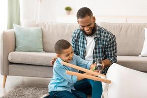 African American Father And Son Fixing Table Together Using Electric Drill Doing House Chores At Home. Happy Kid Helps Dad With Housework Concept. Selective Focus photo