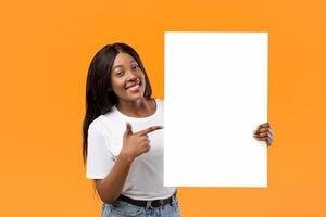 Cheerful african american young lady pointing at empty advertising placard in her hand, orange studio background. Smiling black woman holding blank board for advertisement or text photo