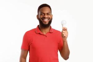 Happy Black Guy Showing Energy-Saving Lightbulb Standing Over White Studio Background, Posing Smiling To Camera. Save Energy, Light Bulb Disposal Service Concept photo