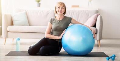 Full length portrait of happy mature woman resting on yoga mat with fitness ball, smiling at camera indoors, free space. Cheerful senior lady taking break from her domestic training photo