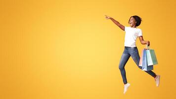 Excited african american young woman with colorful shopping bags jumping in the air and pointing at copy space for sales or discounts advertisement, orange studio background, panorama photo