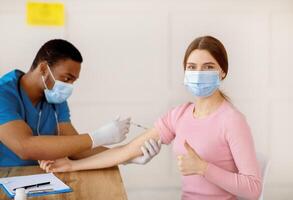 Young woman getting covid-19 vaccine injection, showing thumb up gesture, recommending coromavirus immunization at clinic. Healthcare and viral disease treatment concept photo