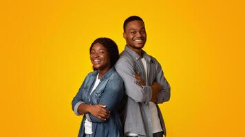 Portrait of confident happy young black man and woman standing back to back with folded arms, cheerful african american couple posing together over yellow background, looking and smiling at camera photo