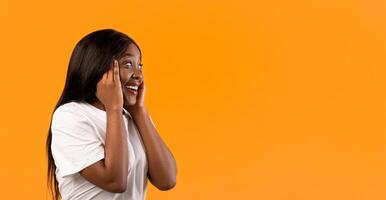 Side view of amazed african american woman with beautiful hair looking at empty space, touching her face in surprised gesture and opening mouth, orange studio background, panorama photo