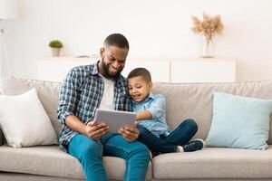 Young Black Father And Little Son Using Tablet Browsing Internet Together And Watching Movie Online Sitting On Sofa At Home On Weekend. Family, Gadgets. Daddy And Child Using Computer Concept photo