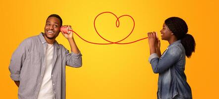 Love Message. Happy black woman whispering romantic words to her boyfriend via tin phone, standing isolated over yellow background, creative collage for Valentine's day with drawn red heart, panorama photo