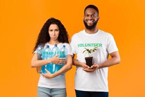 Don't Litter And Care For Nature. Two Volunteers Holding Plant In A Pot And Used Plastic Bottles Standing Over Yellow Studio Background. Ecology, Protection Of Environment Concept photo