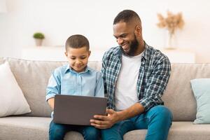 Black Father And Son Using Laptop Computer Watching Cartoons Online Sitting On Sofa Indoors On Weekend. Young Dad And His Kid Browsing Internet Together. Modern Gadgets Concept photo