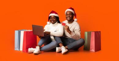 Black Spouses In Santa Hats With Laptop And Credit Card Purchasing Gifts Online, Enjoying Christmas Shopping In Internet, Sitting On Orange Studio Background Next To Bright Paper Shopping Bags photo