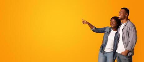 Young Happy Black Couple Pointing With Finger Aside At Copy Space, Excited Young African American Man And Woman Embracing And Looking At Free Place Over Yellow Background, Panorama photo