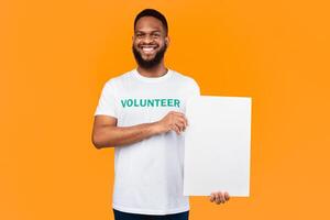 African American Volunteer Man Showing Blank White Paper Board Holding Empty Poster Advertising Your Text Posing Standing Over Yellow Background, Smiling To Camera. Studio Shot, Mockup photo
